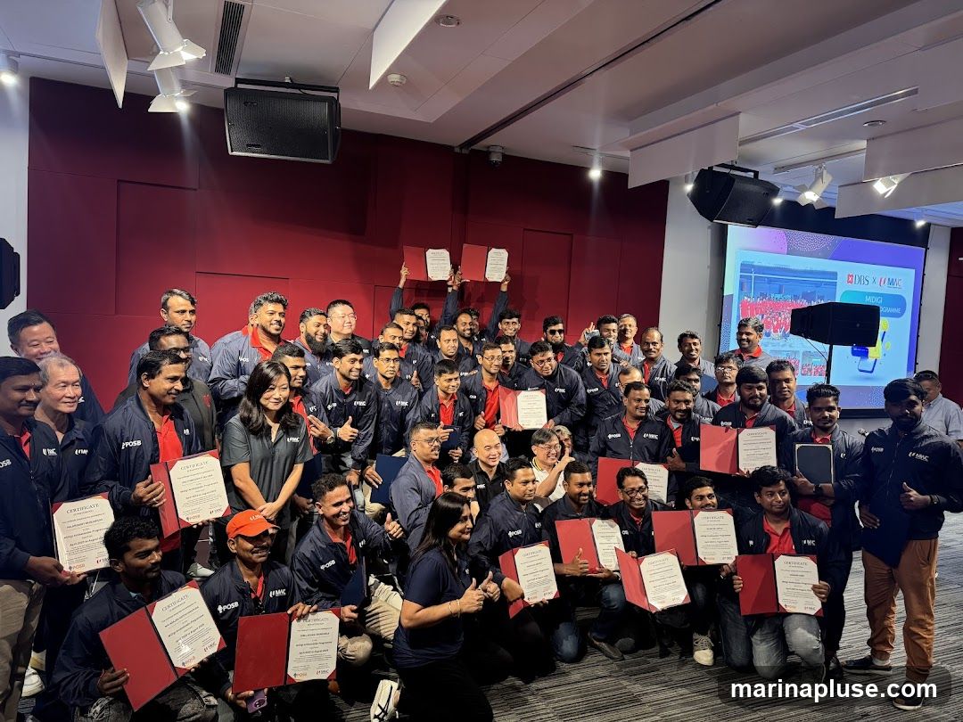 A large group of people, mostly men, are posing for a photo in an indoor setting. Many of them are holding certificates. They appear to be wearing matching dark blue jackets with red undershirts, some with logos visible. A large screen is displayed in the background with text and graphics. The overall atmosphere suggests an awards ceremony or a completion of a program.