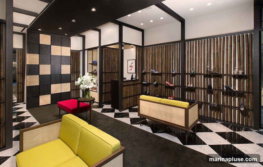 The interior of a modern shoe store features a striking black and light wood checkered wall, bamboo-slatted partitions, and a black and white checkered marble floor. Yellow sofas and a red ottoman provide seating. Shelves display a variety of women's high-heeled shoes. A counter with a framed artwork of shoes is visible in the background.