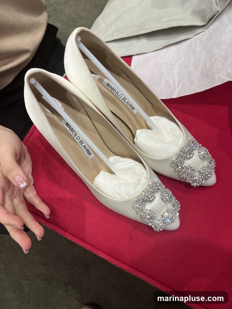 A pair of white satin Manolo Blahnik pumps with crystal embellishments on the toe. The shoes are displayed on a red surface, with a person's hand visible on the left. The brand name "MANOLO BLAHNIK" is visible on labels inside the shoes.