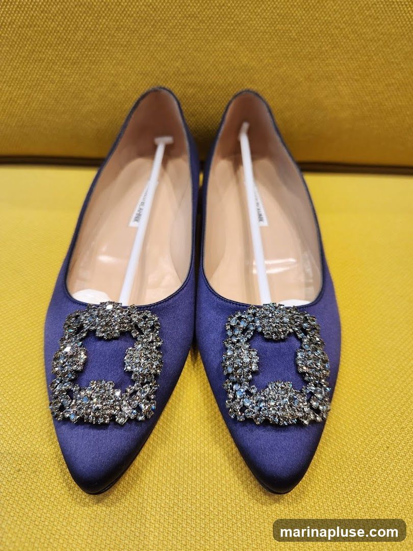 A pair of deep blue satin pointed-toe flats are displayed against a textured yellow background. Each shoe features a large, ornate buckle adorned with numerous clear crystals, placed prominently on the vamp. The interior of the shoes shows a white label with black text, partially visible, reading "MANOLO BLAHNIK". White shoe trees are inserted into each shoe.