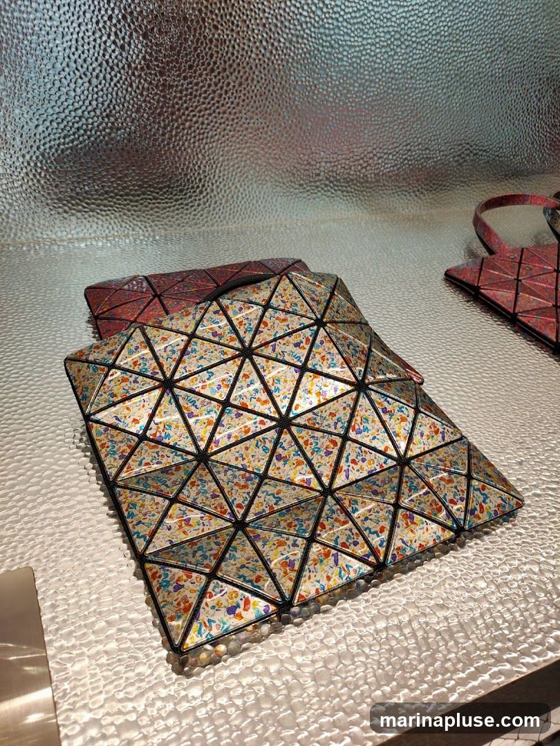 A close-up shot of several geometric-patterned bags displayed on a textured silver surface. The bags are made of triangular panels and feature a colorful confetti-like print. One bag is in the foreground, with two others partially visible behind it. A portion of a pink bag with a handle is visible on the right side of the frame. The background is a reflective, textured silver surface.