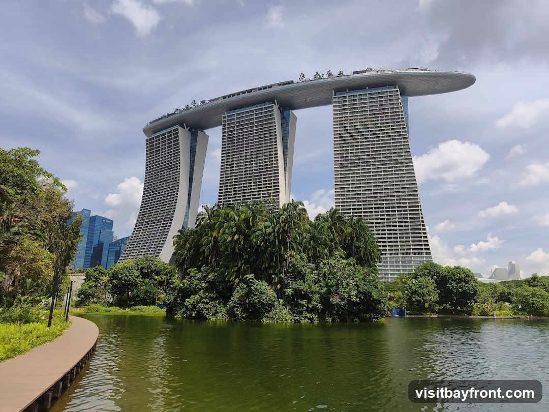 A wide shot of the Marina Bay Sands hotel in Singapore, with its distinctive three towers connected by a large rooftop deck. The hotel is surrounded by lush greenery and a body of water in the foreground. A wooden walkway curves along the edge of the water. The sky is partly cloudy.
