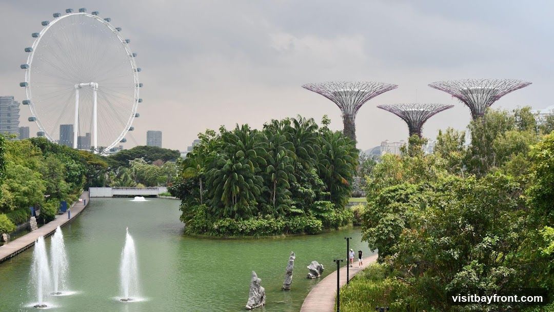 A panoramic view of Gardens by the Bay in Singapore, featuring the Singapore Flyer Ferris wheel in the background, the iconic Supertree Grove, lush greenery, and a tranquil body of water with fountains. People are seen walking along a wooden pathway.
