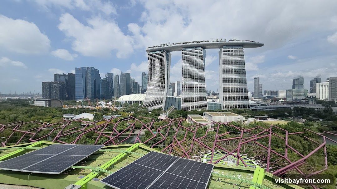 A panoramic view of the Singapore skyline, featuring the iconic Marina Bay Sands hotel, surrounded by numerous skyscrapers. In the foreground, a section of a green structure with solar panels is visible, along with a network of purple-colored metal beams and lush greenery. The sky is partly cloudy.