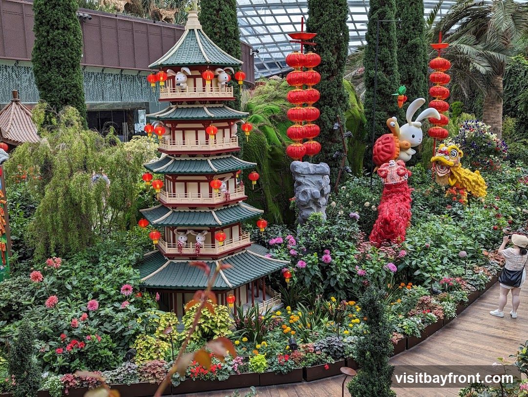 A festive display featuring a multi-tiered pagoda adorned with red lanterns and white rabbits. Nearby, a large white rabbit with a carrot, a turtle, and a yellow lion dance figure are surrounded by lush greenery and colorful flowers. A person is seen taking a photo of the display. The scene appears to be set within a large greenhouse or botanical garden, with a glass ceiling visible in the background.