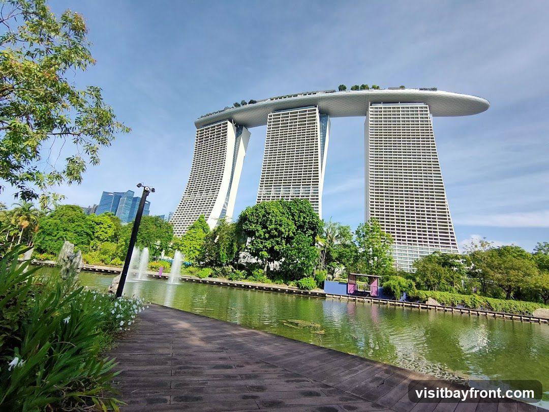 A wide-angle view of the Marina Bay Sands hotel in Singapore, with its distinctive three towers connected by a rooftop deck. The hotel is set against a clear blue sky with scattered clouds. In the foreground, a wooden walkway leads towards a tranquil body of water with fountains. Lush green trees and tropical foliage surround the water, creating a serene park-like atmosphere. The architectural marvel of the hotel is juxtaposed with the natural beauty of the gardens.