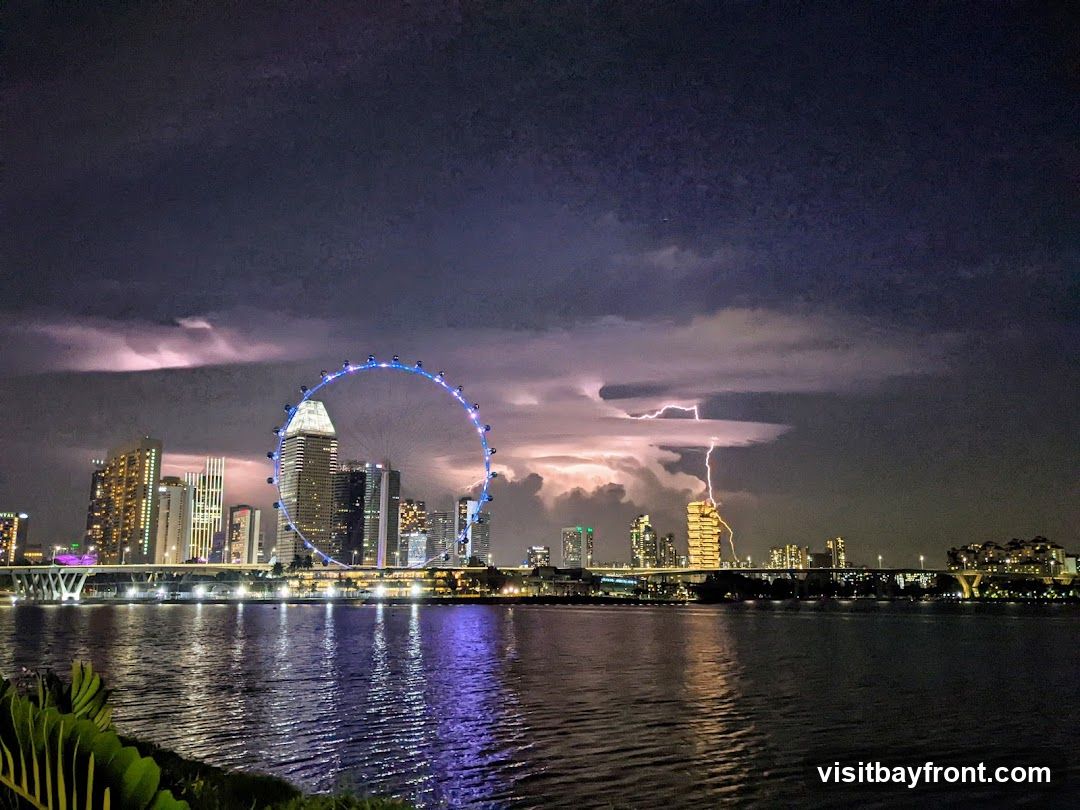 A dramatic nighttime view of the Singapore skyline with lightning striking in the distance. The Singapore Flyer Ferris wheel is illuminated, and city lights reflect on the water in the foreground.