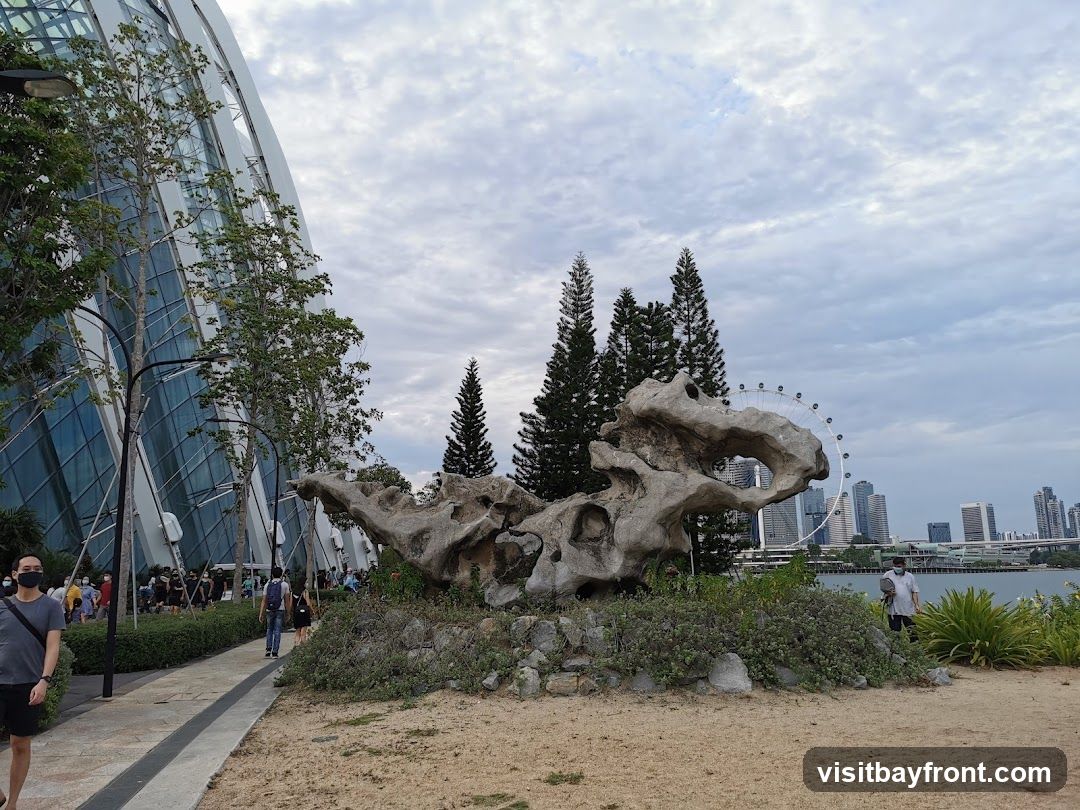 A scenic outdoor view featuring a large, weathered rock formation in the foreground, resembling a mythical creature. Behind it, a cluster of tall pine trees stands against a cloudy sky. In the distance, a Ferris wheel and a modern city skyline are visible across a body of water. To the left, a portion of a large, glass-domed structure, likely a conservatory or exhibition hall, is seen with people walking along a path beside it. The overall scene captures a blend of nature, art, and urban landscape.