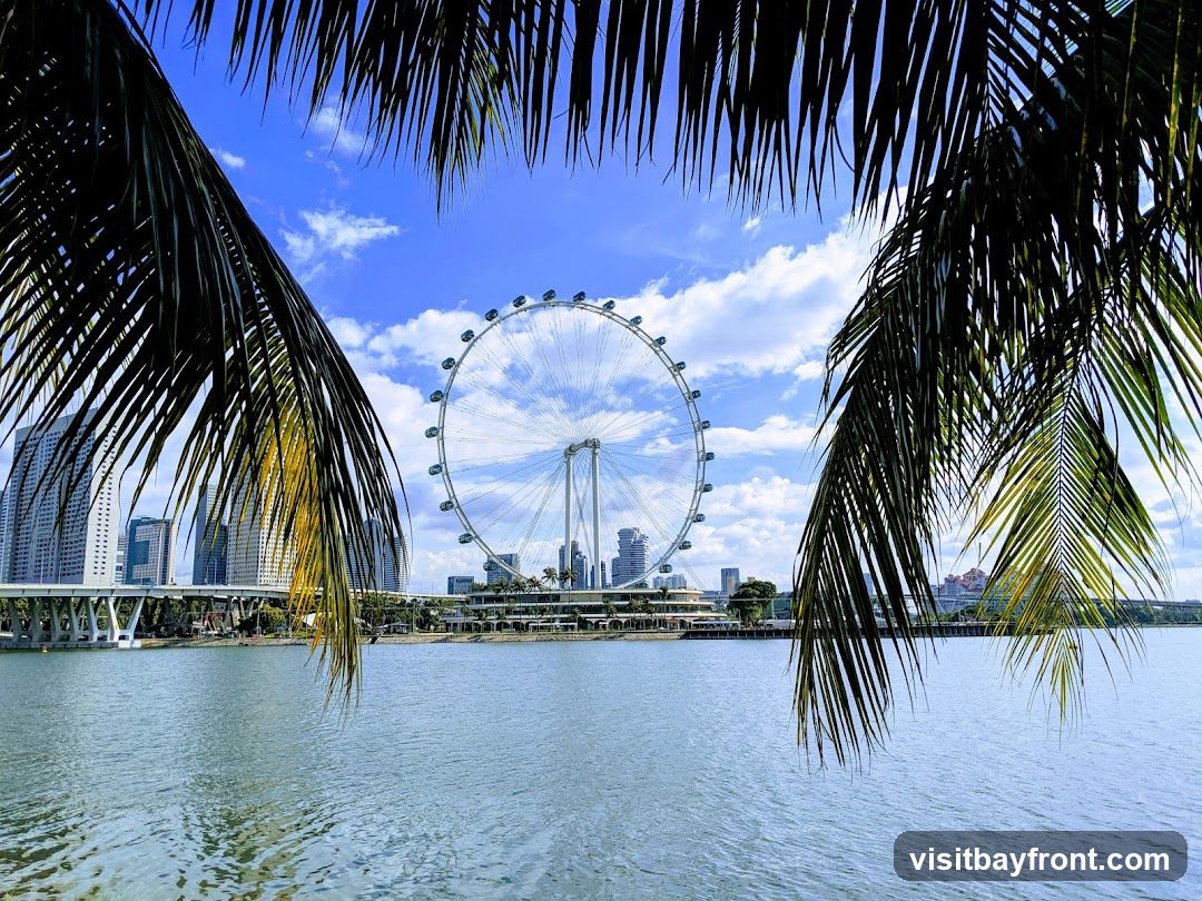 A view of the Singapore Flyer observation wheel and the city skyline across a body of water, framed by palm fronds. The sky is blue with scattered clouds.