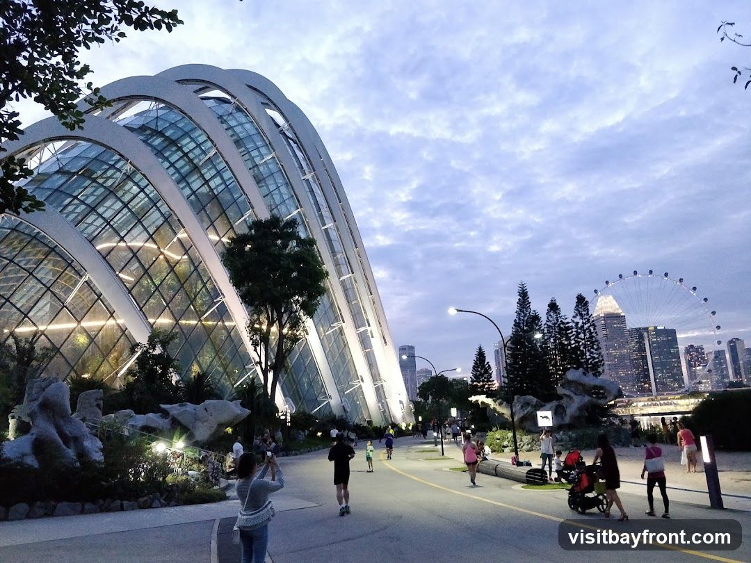 A wide shot of Gardens by the Bay in Singapore during dusk. The iconic Supertree Grove and Cloud Forest domes are visible on the left, illuminated from within. A pathway curves through the park, with several people walking and jogging. In the background, the Singapore skyline and the Singapore Flyer Ferris wheel are visible against a cloudy sky.