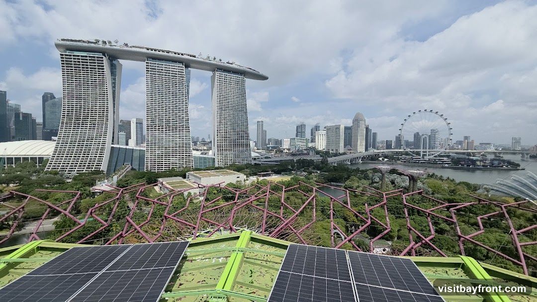 A panoramic view of the Singapore skyline, featuring the iconic Marina Bay Sands hotel with its distinctive ship-like structure on top. In the foreground, solar panels are visible on a green rooftop, overlooking the lush greenery and futuristic structures of Gardens by the Bay. The Singapore Flyer Ferris wheel can be seen in the distance, along with bridges and other skyscrapers, under a partly cloudy sky.
