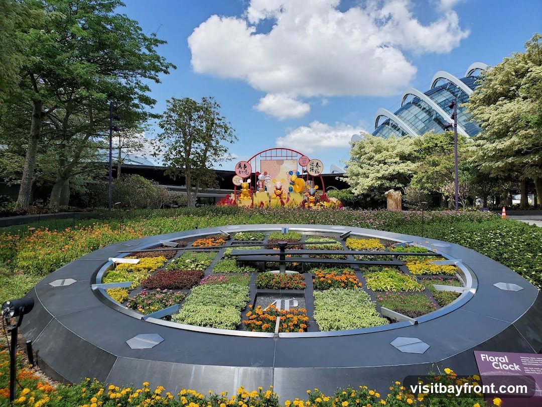 A large, circular floral clock display with various colorful plants arranged in sections. In the background, there is a festive display with cartoon characters and Chinese New Year decorations, set against the backdrop of the iconic domes of Gardens by the Bay. The sky is blue with scattered white clouds.