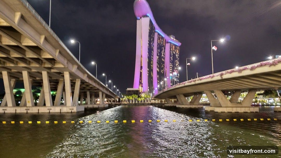 A nighttime view of the Marina Bay Sands hotel in Singapore, illuminated with purple and pink lights. Two elevated highways or bridges flank the scene, with the water of Marina Bay in the foreground. Yellow buoys mark a boundary in the water.