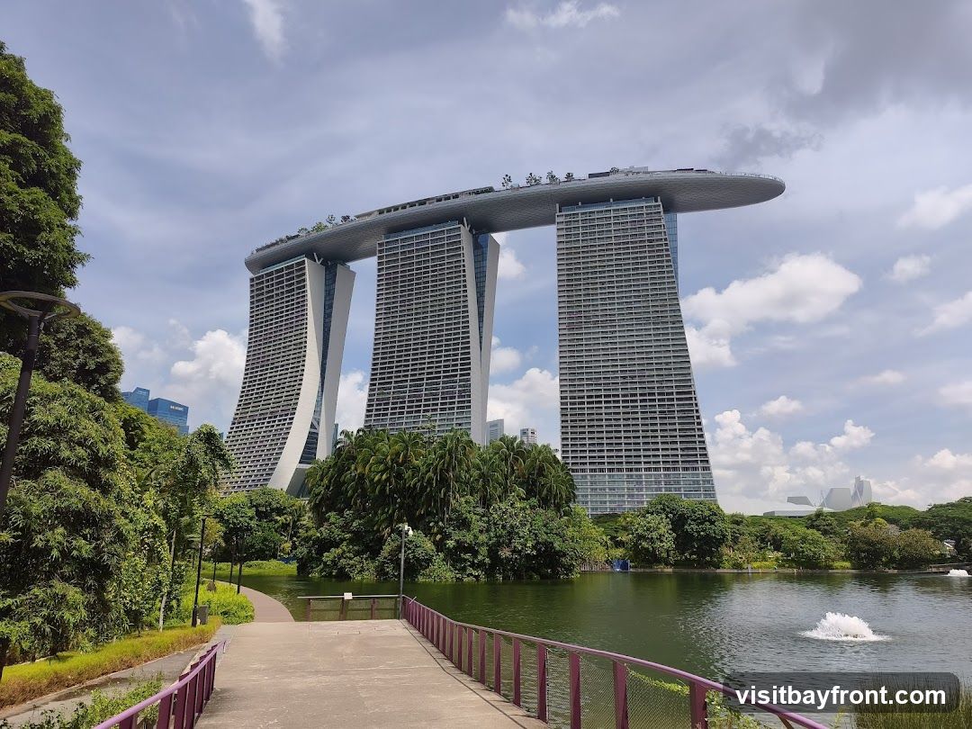 A wide shot of the Marina Bay Sands hotel in Singapore, with its distinctive three towers connected by a large, flat roof structure. The hotel is set against a cloudy blue sky. In the foreground, there is a lush green park with a body of water, a pathway, and trees. A fountain is visible in the water.