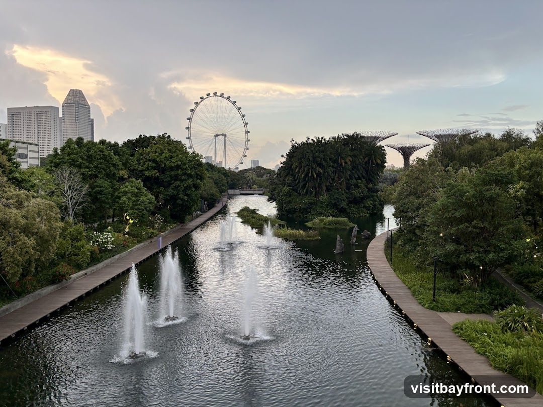 A scenic view of a park with a canal featuring multiple fountains. A wooden walkway lines the canal, with lush greenery and trees on either side. In the background, a Ferris wheel and distinctive futuristic tree-like structures are visible, along with a city skyline featuring modern skyscrapers. The sky is overcast with soft, diffused light.
