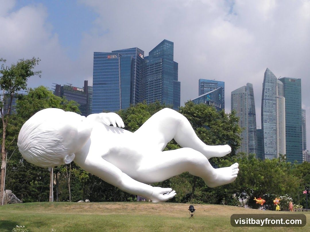 A large, white sculpture of a sleeping baby floats in the foreground, set against a backdrop of modern skyscrapers and lush green trees under a partly cloudy sky. The sculpture is detailed, showing the baby's head, torso, arms, and legs. In the distance, several tall buildings with glass facades, including those with 'DBS' and 'MUFG' logos, dominate the skyline. The foreground features a grassy lawn with some small plants and a spotlight on the ground.