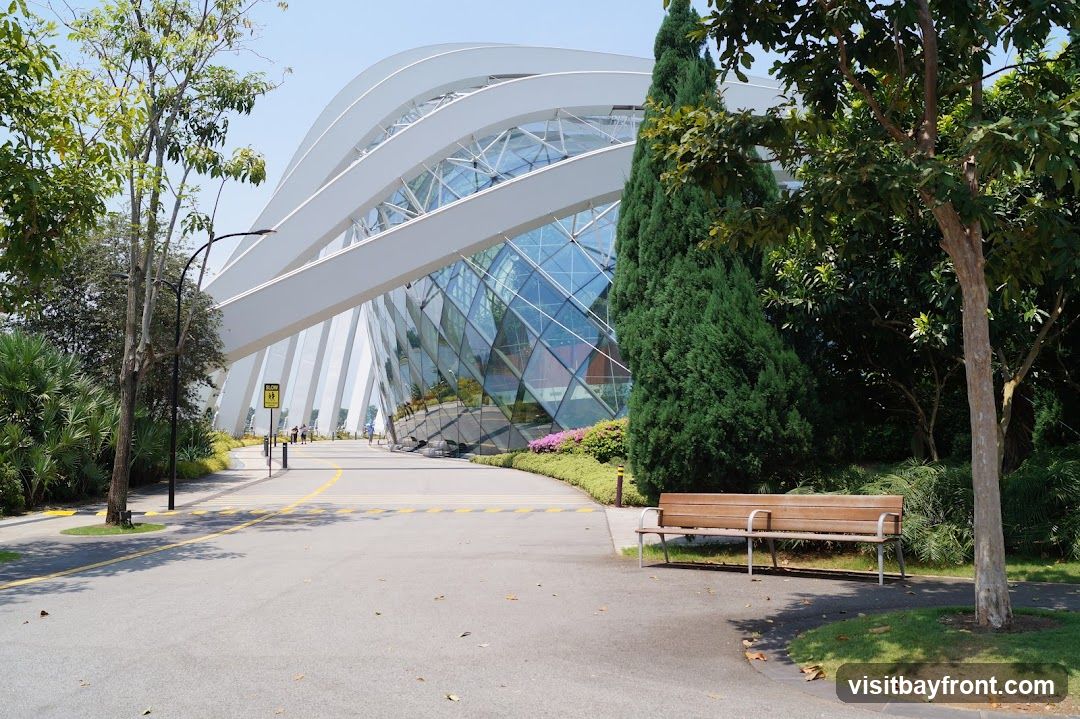 A wide shot of the exterior of the Gardens by the Bay conservatory, with its distinctive white, curved roof and glass facade. A paved pathway leads towards the entrance, with lush greenery and trees on either side. A wooden bench is visible in the foreground. Several people are walking on the path in the distance.