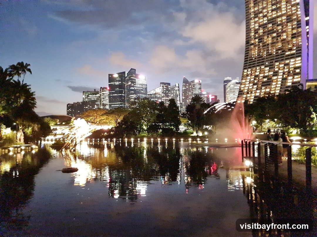 A nighttime view of a city skyline with illuminated skyscrapers reflected in a body of water. In the foreground, a fountain sprays pink water, and lights line the edge of the water. Palm trees and other foliage are visible on the left, and people can be seen standing near the fountain on the right. The sky is a deep blue with scattered clouds.