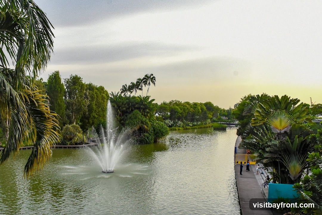 A scenic view of a park with a large body of water featuring a central fountain. Lush green trees and palm trees line the water's edge, with a paved walkway visible on the right side where a few people are standing. The sky is overcast with a hint of sunset glow.