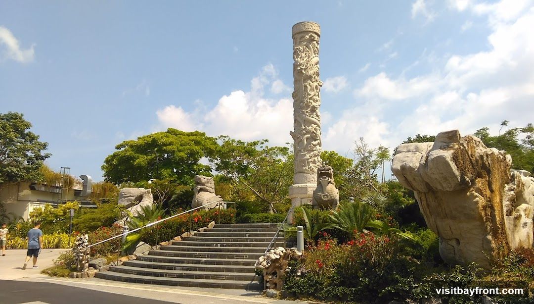 A tall, intricately carved stone pillar stands in a lush garden setting. The pillar is adorned with detailed carvings of dragons and other mythical creatures. In front of the pillar, a wide set of stone stairs leads up into the garden. Flanking the stairs are two stone lion statues. The garden features a variety of plants, including palm trees, flowering bushes, and large rocks. In the background, there are trees and a building with visible air conditioning units. Two people are walking on a paved path to the left of the stairs. The sky is blue with scattered white clouds.
