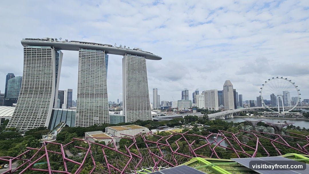 A panoramic view of the Singapore skyline, featuring the iconic Marina Bay Sands hotel with its distinctive ship-like structure connecting three towers. In the background, the Singapore Flyer Ferris wheel is visible, along with other skyscrapers and bridges. The foreground shows a section of a green structure with pink lattice work and solar panels, suggesting a viewpoint from a garden or elevated platform. The sky is partly cloudy.