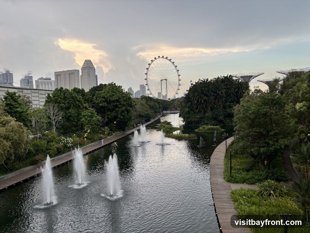 A scenic view of a park with a body of water featuring fountains. A wooden walkway lines the right side of the water. In the background, a city skyline with tall buildings and a large Ferris wheel is visible under a cloudy sky. The distinctive Supertree Grove structures of Gardens by the Bay can also be seen on the right side of the image.