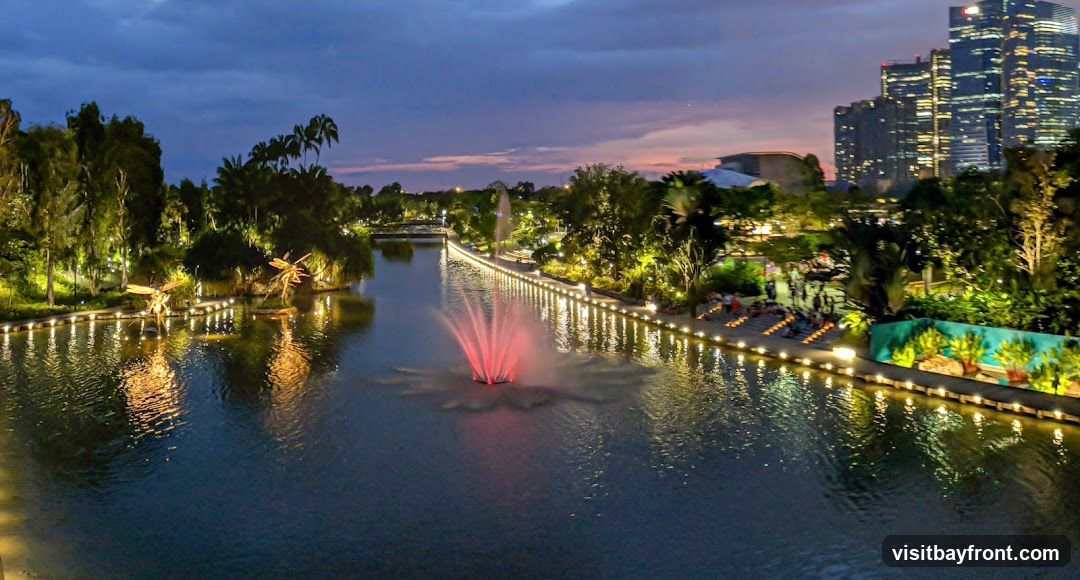 A panoramic view of Gardens by the Bay in Singapore at dusk. A central body of water features a fountain with red lights. The water is illuminated by lights along the pathways and reflects the surrounding lush greenery and palm trees. In the background, modern skyscrapers glow against the twilight sky. People are visible along the illuminated walkways, enjoying the evening ambiance.