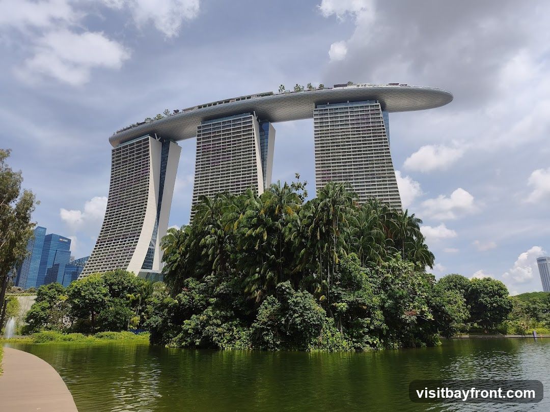 A wide shot of the Marina Bay Sands hotel in Singapore, with its iconic three towers connected by a rooftop deck. The hotel is surrounded by lush greenery and a body of water in the foreground. The sky is partly cloudy.