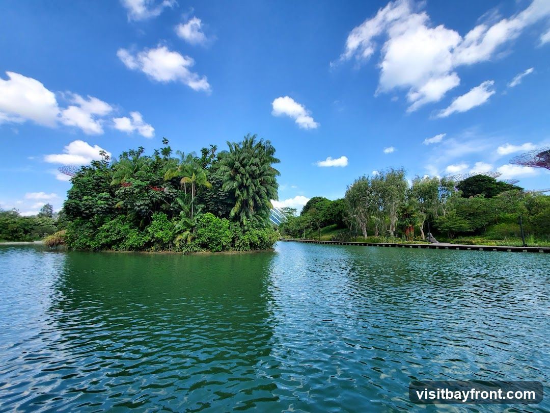 A serene landscape featuring a vibrant green island covered in dense foliage and palm trees, situated in the middle of a tranquil, blue-green lake. The sky above is a brilliant blue, dotted with fluffy white clouds. A wooden boardwalk runs along the far bank of the lake, lined with various trees and some futuristic structures in the distance. The water's surface is gently rippled, reflecting the sky and surrounding greenery.
