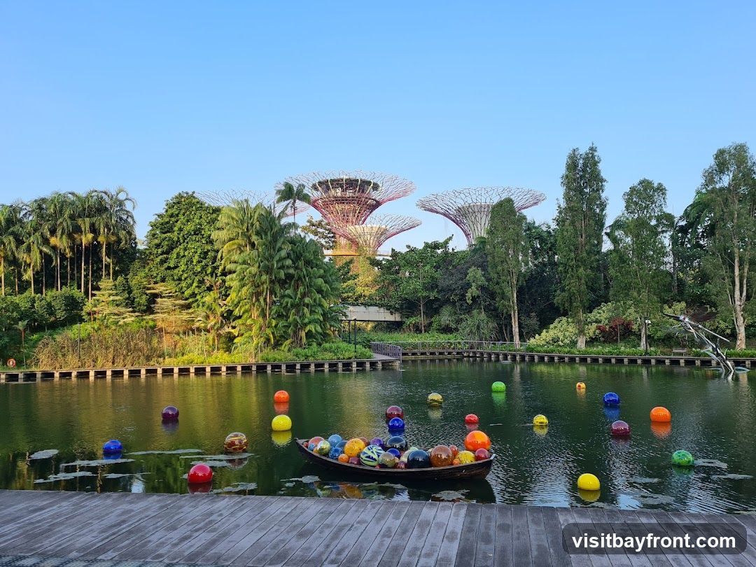 A scenic view of Gardens by the Bay in Singapore, featuring the iconic Supertree Grove in the background. A calm body of water in the foreground is adorned with colorful glass spheres floating on its surface, with a small boat filled with more spheres docked on the right. Lush greenery and palm trees surround the water, creating a vibrant and serene landscape under a clear blue sky.
