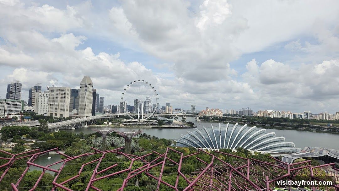 A panoramic view of the Singapore skyline, featuring the Marina Bay Sands, Singapore Flyer, and the iconic Supertree Grove and Cloud Forest domes of Gardens by the Bay. The cityscape is set against a backdrop of a cloudy sky.
