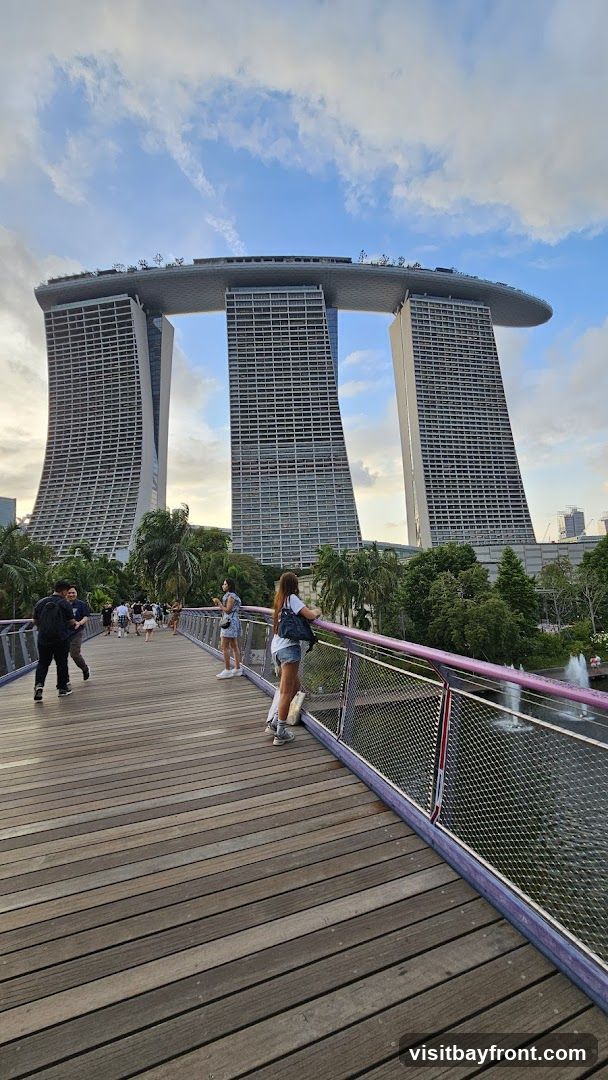 A wide shot of the Marina Bay Sands Hotel in Singapore, with people walking on a wooden bridge in the foreground. The sky is partly cloudy with blue patches.