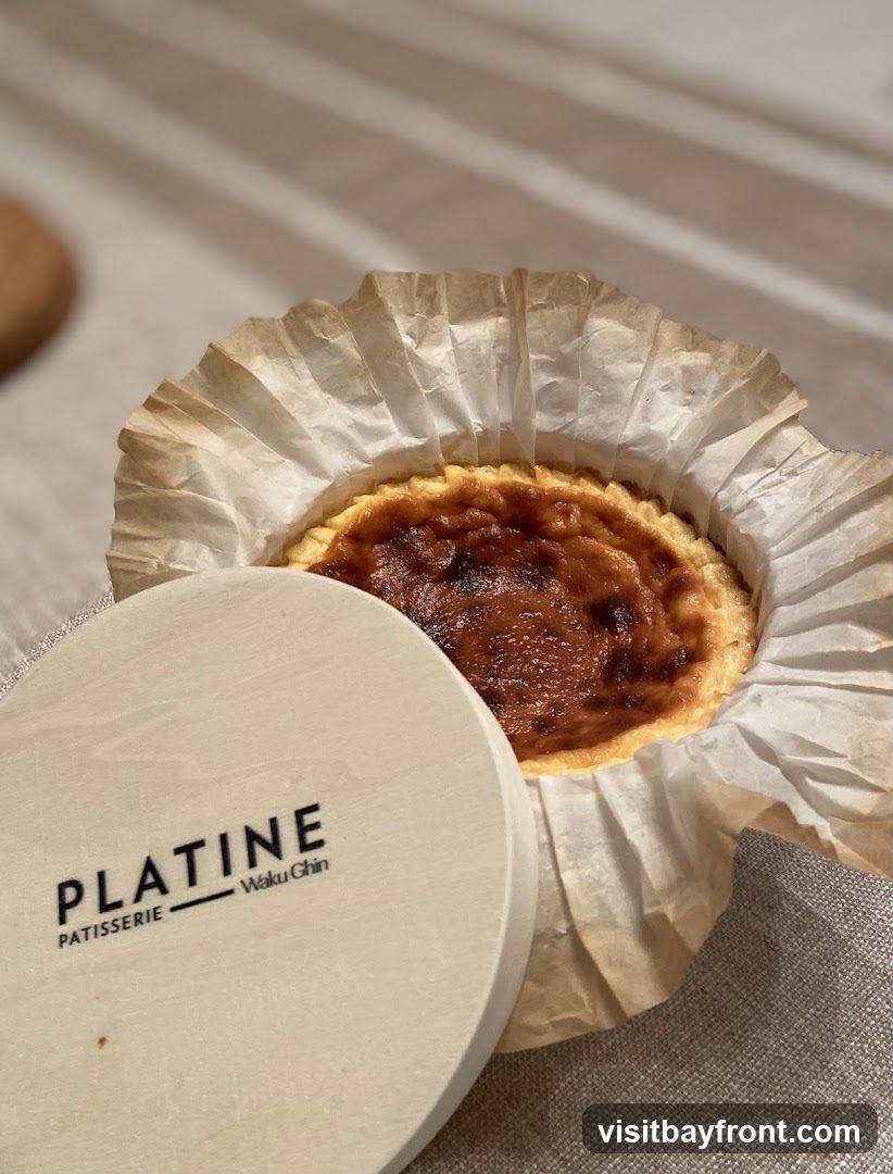 A close-up of a Burnt Basque cheesecake, presented in a paper liner and partially covered by a wooden lid. The lid is branded with "PLATINE PATISSERIE - Waku Ghin". The cheesecake has a caramelized, slightly charred top and a creamy interior visible around the edges. The setting appears to be a table with a textured tablecloth.