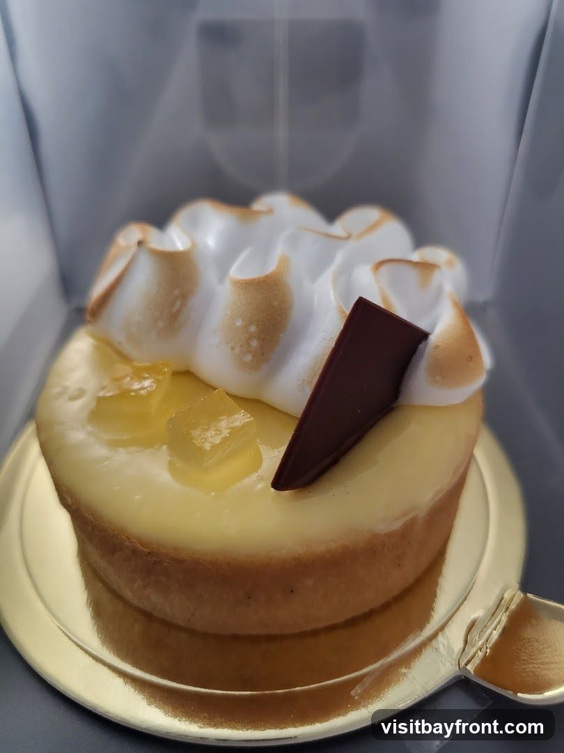 A close-up shot of a lemon meringue tart. The tart has a golden crust, filled with a smooth yellow lemon curd, topped with torched meringue and garnished with small cubes of jelly and a dark chocolate shard. The tart is presented on a gold cake board within a white box.