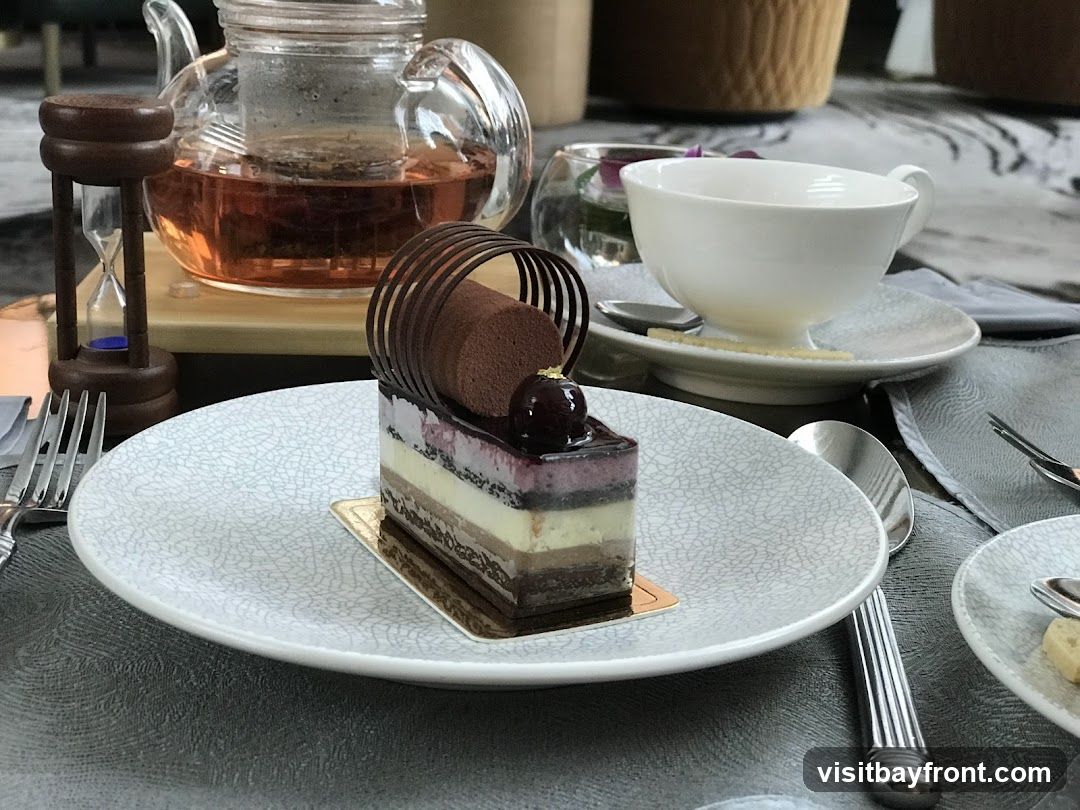 A slice of layered chocolate cake with a cherry on top sits on a white plate with a textured pattern. A glass teapot filled with amber tea and a tea infuser is in the background, along with a white teacup and saucer. A small hourglass timer is also visible.