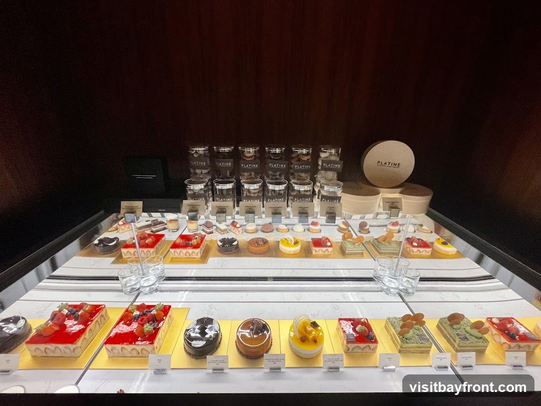 A display case filled with various pastries and desserts. The desserts include tarts, cakes, and small individual pastries, many topped with fruits like strawberries and raspberries. Some items are arranged in rectangular and circular shapes. Branded packaging with the name 'PLATINE' is visible in the background.