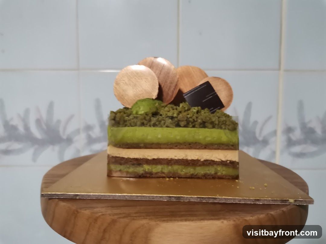 A slice of matcha layer cake is presented on a golden square board, which rests on a wooden stand. The cake features distinct layers of green and light brown mousse, with a dark brown base. The top is adorned with crumbled matcha cake, small green spheres, wooden discs, and a dark chocolate square. The background consists of light blue tiles with a subtle shadow pattern.