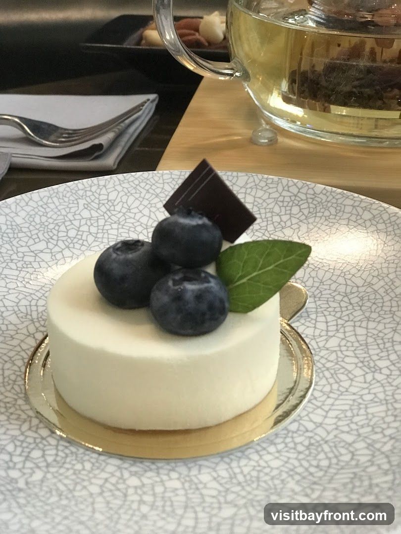 A close-up of a small, white dessert topped with blueberries, a piece of dark chocolate, and a mint leaf. The dessert is on a patterned plate. In the background, a glass teapot filled with tea and a small dish of nuts are visible, along with a fork and napkin.