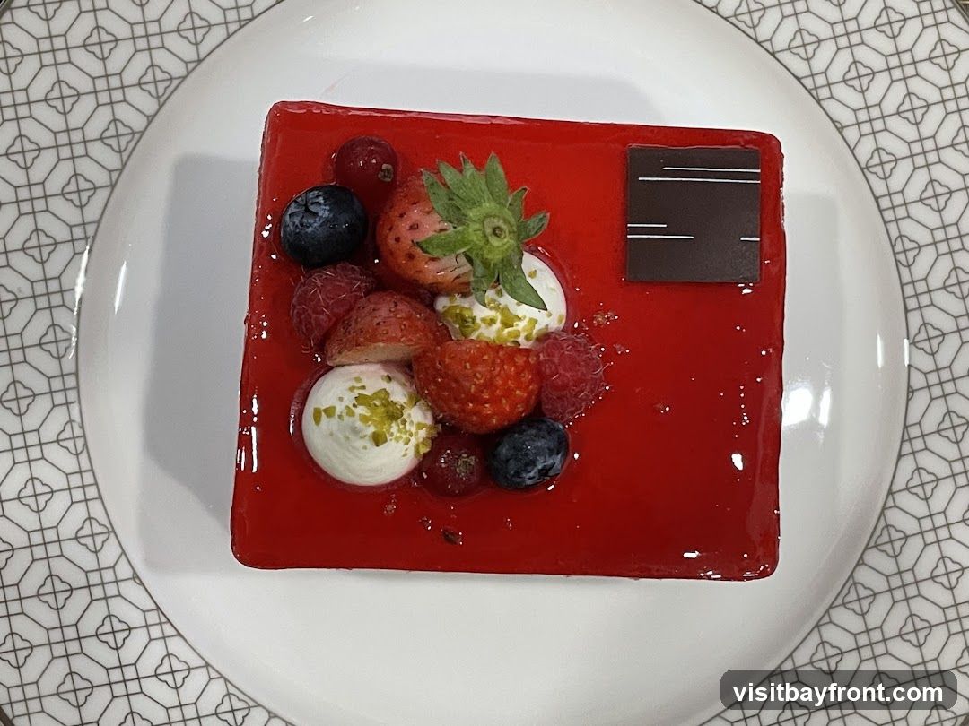 A square red berry mousse cake topped with fresh strawberries, blueberries, raspberries, red currants, dollops of whipped cream, and chopped pistachios. A dark chocolate square with white lines is placed on the top right corner of the cake. The cake is served on a white plate with a geometric pattern around the rim.
