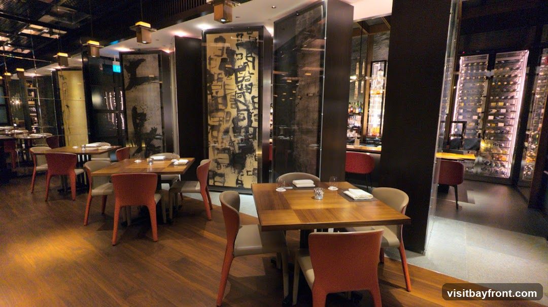 The image shows the interior of a modern restaurant. Tables are set with white napkins and glassware. The decor includes large abstract art panels and illuminated wine displays. The flooring is polished wood, and the lighting is ambient.
