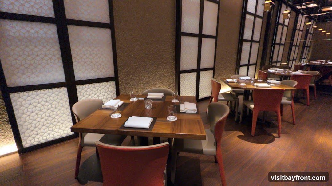 A well-lit dining area in a restaurant with multiple tables set for guests. The tables are made of dark wood and are set with white napkins, glasses, and cutlery. The chairs are a mix of beige and red. The walls are textured in a warm brown tone and feature decorative panels with a geometric pattern. The flooring is polished wood.