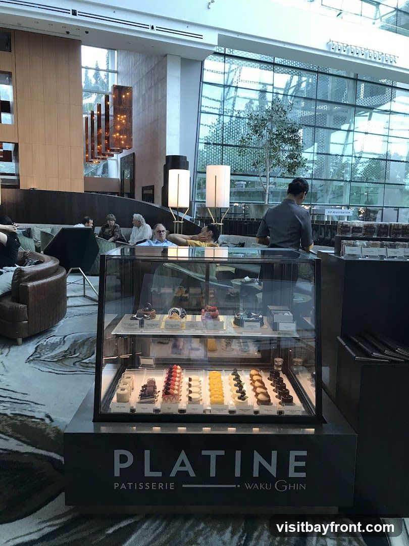A display case filled with various pastries and desserts is shown in a well-lit interior space. The display case has two shelves, each showcasing rows of meticulously arranged treats. The bottom shelf features items like eclairs, tarts, and small cakes, while the top shelf displays more elaborate cakes and pastries. The front of the display case has a prominent sign that reads "PLATINE PATISSERIE WAKU GHIN". In the background, people are seated in a lounge area, and a large window offers a view of the outdoors and modern architecture.