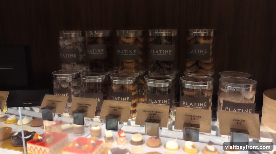 A display of various pastries and cookies in clear plastic containers, with labels indicating different flavors and prices. The brand name 'PLATINE' is visible on the containers.