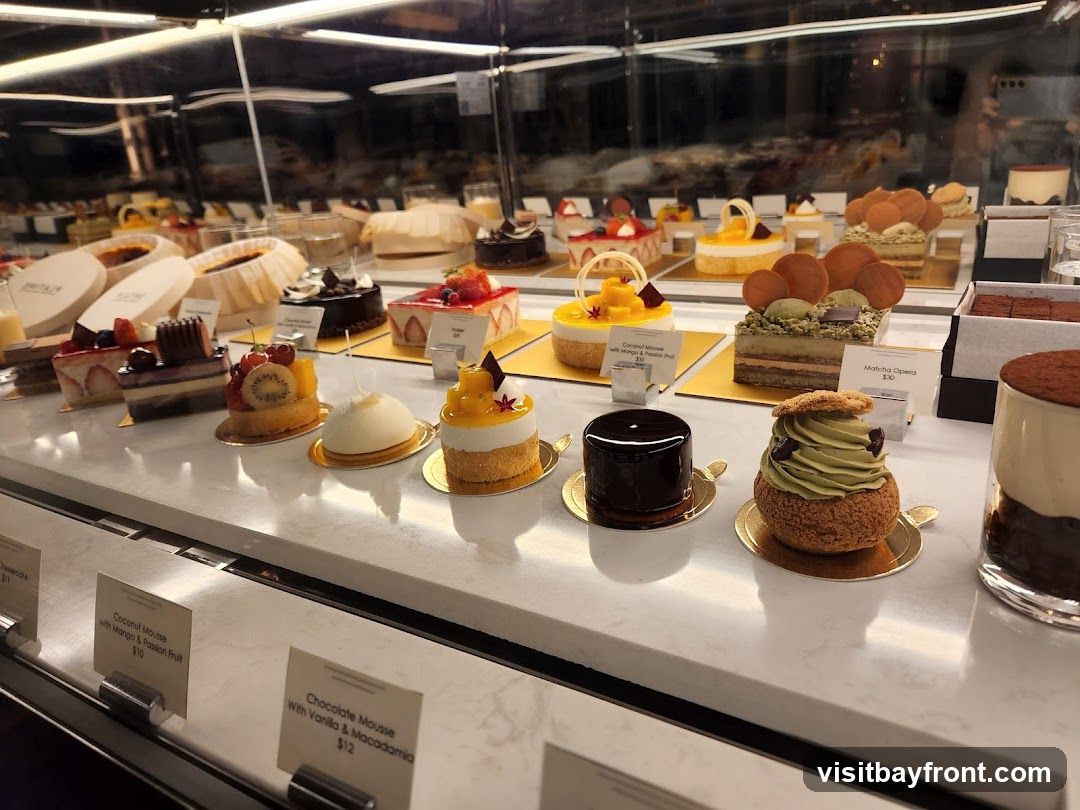 A display case filled with various colorful and elaborately decorated pastries and cakes. In the foreground, there are several individual desserts, including a dark chocolate mousse, a matcha cream puff, and a layered cake. Labels indicate prices for some items: Coconut Mousse with Mango & Passion Fruit ($10), Chocolate Mousse with Vanilla & Macadamia ($12), and Matcha Opera ($30). In the background, more desserts are visible, including tarts and cakes, some in boxes.