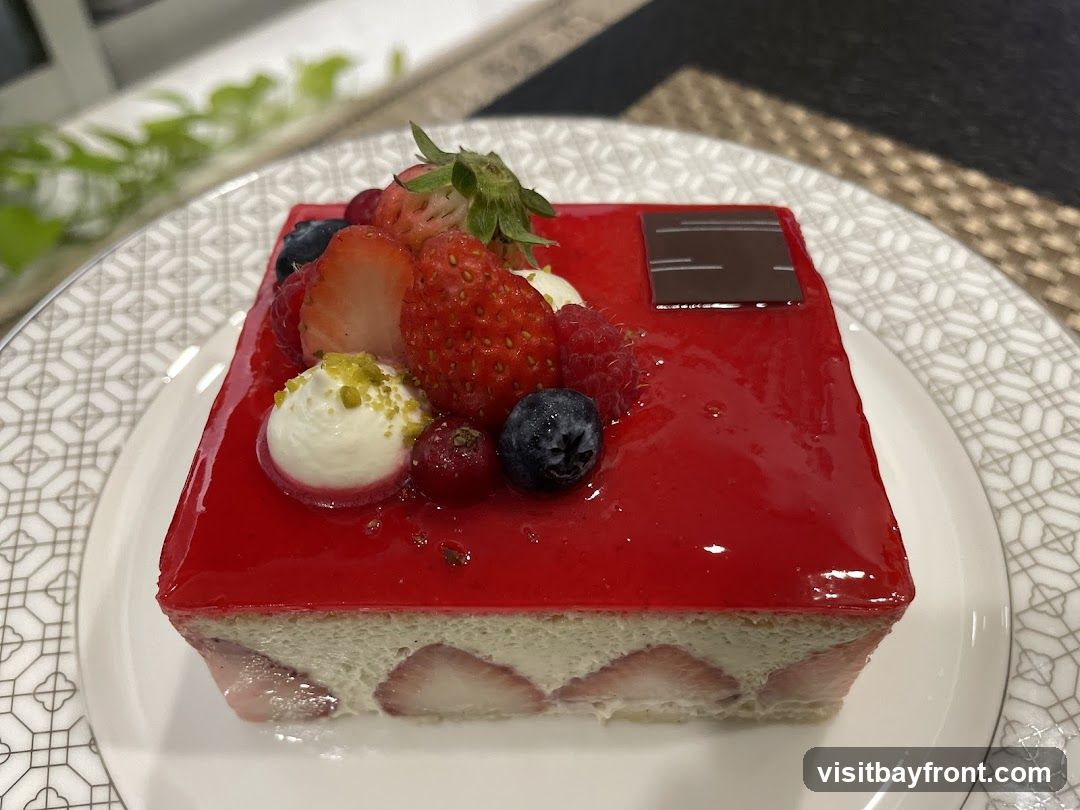 A slice of strawberry mousse cake topped with fresh berries and a chocolate square. The cake has a glossy red glaze and a creamy white mousse layer with visible strawberry slices. It is served on a white plate with a decorative pattern.