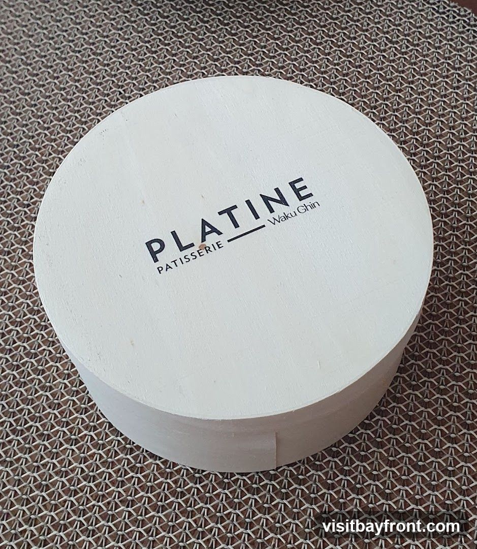 A white, round cake box with the words "PLATINE PATISSERIE" and "Waku Chin" printed on the lid. The box is placed on a textured brown surface.