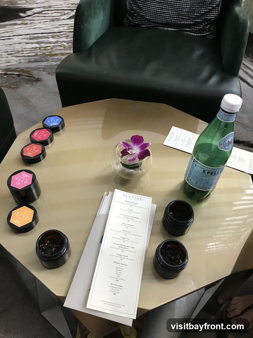 A table is set with several small containers of loose leaf tea, a bottle of San Pellegrino water, and a menu. A small orchid in a glass vase adds a decorative touch. The setting appears to be in a lounge or dining area with plush seating visible in the background.