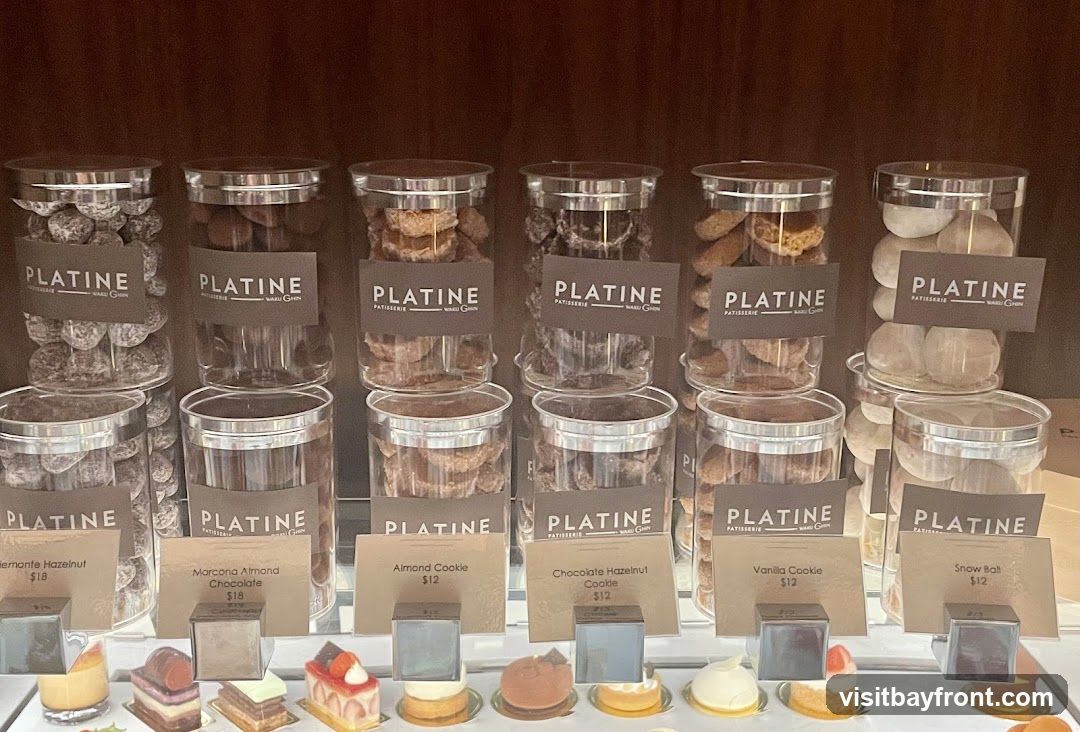 A display case filled with various pastries and cookies in clear jars, labeled with the brand "PLATINE PATISSERIE". Below the jars are small cards detailing specific items and their prices. Some of the items include "Hermonte Hazelnut" for $18, "Marcona Almond Chocolate" for $18, "Almond Cookie" for $12, "Chocolate Hazelnut Cookie" for $12, "Vanilla Cookie" for $12, and "Snow Ball" for $12.