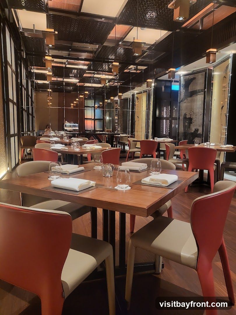 A well-lit, modern dining room with multiple tables set for guests. The tables are made of wood and feature white napkins, silverware, and glassware. The chairs are a mix of beige and red. The room has a reflective ceiling with pendant lights and decorative wall panels.