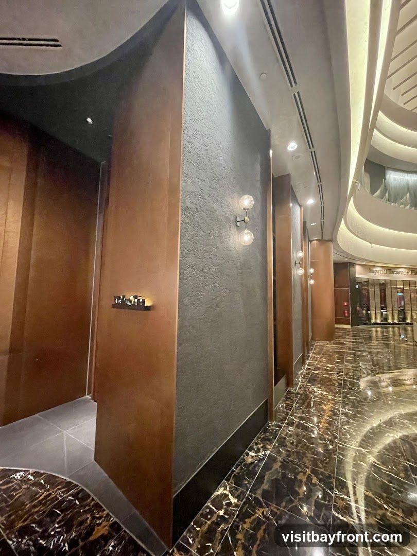 A view down an interior hallway with dark marble flooring and textured walls. There are modern light fixtures on the walls and ceiling. In the distance, a sign for "Imperial Treasure Fine Chinese Cuisine" is visible.